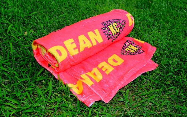 May include: A pink beach towel with the name "DEAN" written in yellow letters. The towel has a black and yellow leopard print arrow design with the letters "KC" in the center.