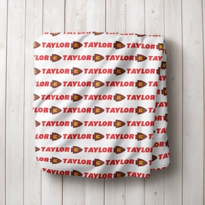 May include: White fabric with a repeating pattern of red "TAYLOR" text with a red and yellow arrow and "KC" in a yellow circle.