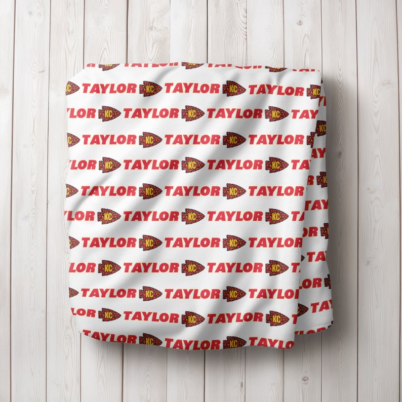 May include: White fabric with a repeating pattern of red "TAYLOR" text with a red and yellow arrow and "KC" in a yellow circle.