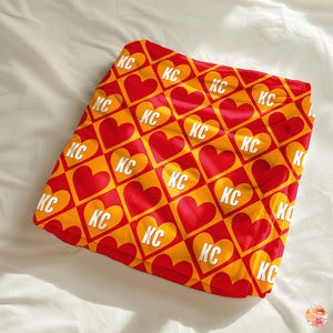 May include: A red and yellow throw blanket with a repeating pattern of hearts and the letters "KC".