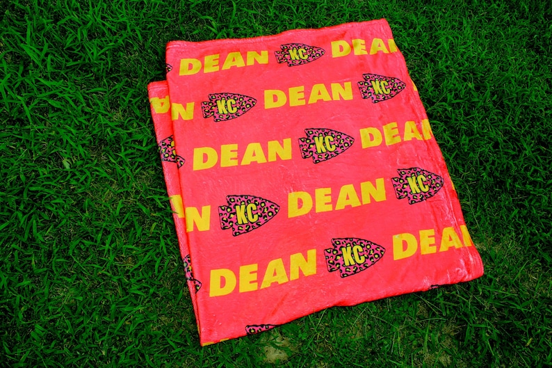 May include: A pink fleece blanket with a repeating pattern of yellow "DEAN" text and pink and black leopard print arrowheads with "KC" inside.