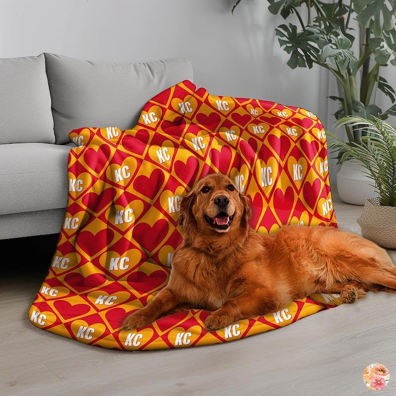 May include: A red and yellow fleece blanket with a repeating pattern of hearts and the letters "KC". A golden retriever dog is lying on the blanket.