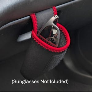 May include: Black sunglasses holder with red edging, designed for car interiors. The holder is made of a soft, felt-like material. The image includes the text "(Sunglasses Not Included)".