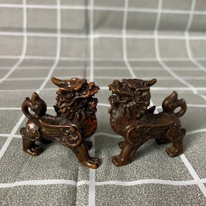 Puede incluir: Dos estatuas de bronce, intrincadamente detalladas, de criaturas míticas. Cada estatua presenta un cuerpo de león con alas, cuernos y una cabeza de dragón. Las estatuas están posadas en direcciones opuestas, mostrando su detallada artesanía.