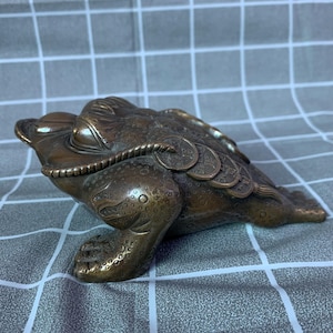 May include: Bronze-colored frog sculpture with coins on its back. The frog has a textured skin and detailed features, including a rope-like collar. The sculpture is a decorative item, likely for home decor or as a collectible.