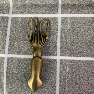 Könnte beinhalten: Ein bronzefarbener Metallverschluss in Tintenfischform. Der Verschluss hat oben zwei gebogene Schlaufen, ein detailliertes Tentakeldesign und einen sich verjüngenden Körper. Der Verschluss dient zum Sichern von Gegenständen.