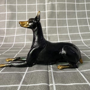 May include: A black and gold figurine of a Doberman Pinscher dog in a lying down position. The sculpture features a glossy black body with metallic gold accents on the snout, the tips of the ears, and the paws, capturing the alert posture.