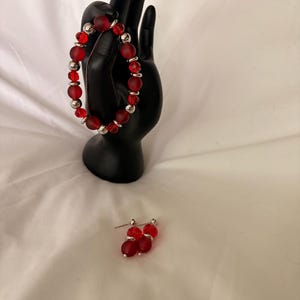 May include: A red beaded bracelet and matching earrings. The bracelet features frosted red beads interspersed with silver-colored metal beads. The earrings have a similar design, with red beads and silver posts. The jewelry is displayed on a black hand-shaped stand.