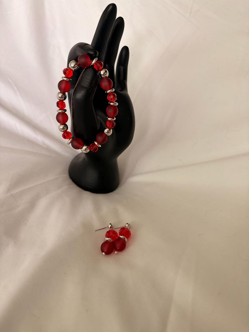May include: A red beaded bracelet and matching earrings. The bracelet features frosted red beads interspersed with silver-colored metal beads. The earrings have a similar design, with red beads and silver posts. The jewelry is displayed on a black hand-shaped stand.