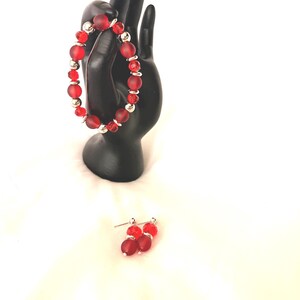 May include: A red beaded bracelet and matching earrings set. The bracelet features large, frosted red beads interspersed with smaller, clear red beads and silver-colored accents. The earrings have a similar design, with red beads and silver posts.