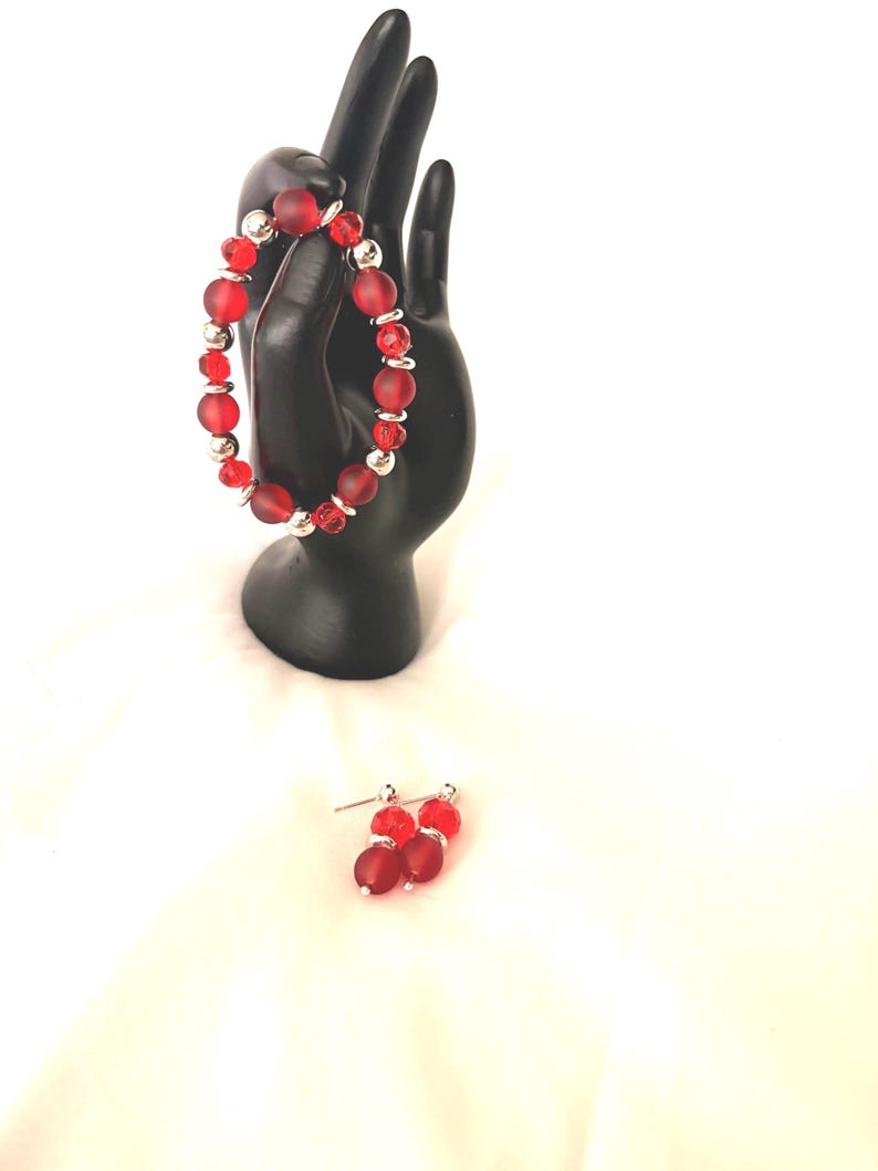 May include: A red beaded bracelet and matching earrings set. The bracelet features large, frosted red beads interspersed with smaller, clear red beads and silver-colored accents. The earrings have a similar design, with red beads and silver posts.