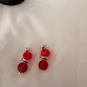 May include: A pair of red beaded earrings. Each earring features a small silver stud, a faceted red bead, a silver disc, and a larger, matte red bead. The earrings are displayed on a white surface.