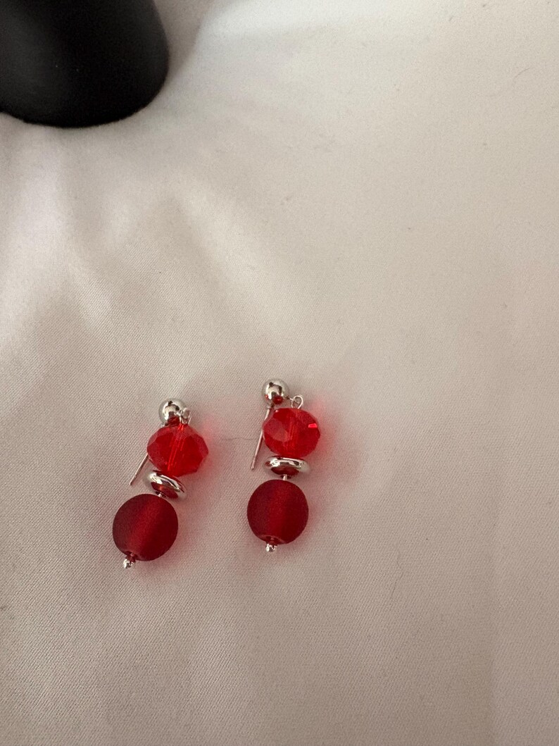 May include: A pair of red beaded earrings. Each earring features a small silver stud, a faceted red bead, a silver disc, and a larger, matte red bead. The earrings are displayed on a white surface.