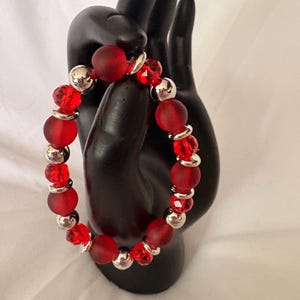 May include: A red beaded bracelet with a mix of matte and faceted beads. The bracelet features large, round, matte red beads interspersed with smaller, faceted red beads and silver-colored metal beads. The bracelet is displayed on a black hand.