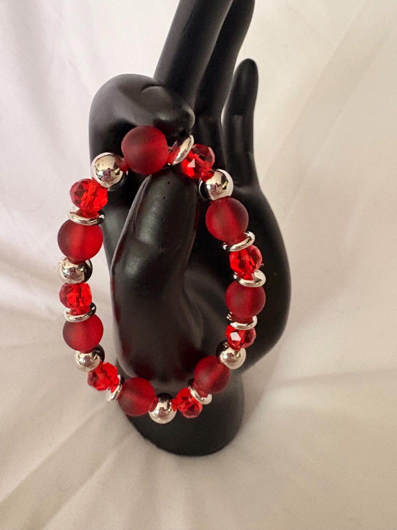 May include: A red beaded bracelet with a mix of matte and faceted beads. The bracelet features large, round, matte red beads interspersed with smaller, faceted red beads and silver-colored metal beads. The bracelet is displayed on a black hand.
