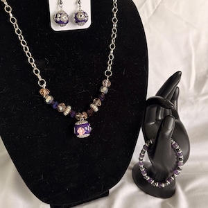 May include: A jewelry set featuring a necklace, earrings, and bracelet. The necklace has a silver chain with purple and clear beads, and a purple pendant with a floral design. The earrings and bracelet have matching purple beads. The jewelry is displayed on a black stand.