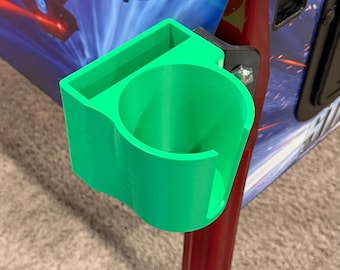 May include: A bright green 3D-printed cup holder attached to a red pinball machine leg. The cup holder is cylindrical with a rectangular top, designed to hold a beverage. The pinball machine has a blue and red graphic.