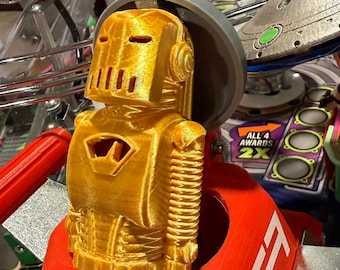 May include: A gold-colored robot figurine with a helmet and chest emblem, set on a red base. The robot is part of a pinball machine, with other elements like a spaceship and text reading "Star Fighter Launch" visible in the background.