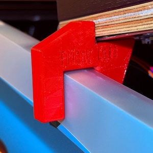 以下が含まれることがあります： 赤色の3Dプリントされたブラケットのクローズアップ。側面には「FUNHOUSE」の文字があり、水色とシルバーの表面に取り付けられています。ブラケットは、電子部品とワイヤーを備えた木製パネルを支えています。