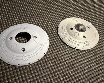 May include: Two circular, metallic-looking objects with a futuristic design. One is white with a central hole and circular patterns. The other is silver with a central, textured cone and small holes.