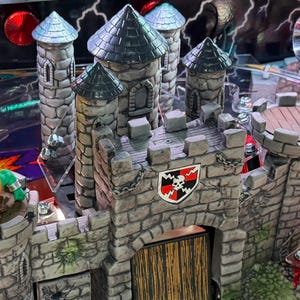 May include: A detailed view of a pinball machine with a castle theme. The castle is made of gray stone with a shield bearing a skull and crossbones. A green monster figure is on a tower. The machine has a clear plastic top.
