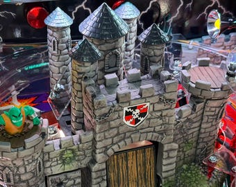 May include: A detailed view of a pinball machine with a castle theme. The castle is made of gray stone with a shield bearing a skull and crossbones. A green monster figure is on a tower. The machine has a clear plastic top.