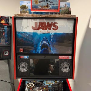 May include: A Jaws-themed pinball machine with a red frame. The backboard features the movie title and a shark image. The top has an "Amity Island" sign. The playfield is visible below, with various game elements.