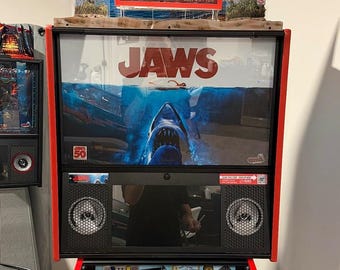 May include: A Jaws-themed pinball machine with a red frame. The backboard features the movie title and a shark image. The top has an "Amity Island" sign. The playfield is visible below, with various game elements.