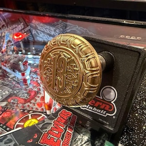 May include: A gold-coloured pinball machine knob featuring the number 13. The knob is mounted on a black surface, with a green button nearby. The words "Stern Pinball" are also visible.