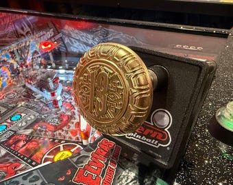May include: Close-up of a gold-colored pinball machine knob with the number 13 in the center. The knob is on a black surface with a green button and the words "Stern Pinball" visible.