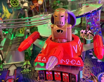 May include: A close-up of a pinball machine with a red and gold robot figure at the center. The robot has a gold head and a red body with the letters "V" and "EA" on it. The pinball machine has green and blue accents.