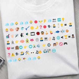 May include: White t-shirt featuring a collection of colorful emoji graphics. The design includes various icons such as faces, objects, and symbols, arranged in rows across the front of the shirt. The shirt is made of a soft, comfortable material.