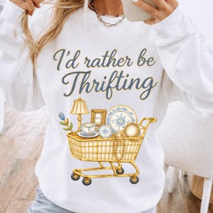 May include: White sweatshirt with the phrase "I'd rather be Thrifting" in blue script. A gold shopping cart graphic is filled with vintage items like a lamp, teacup, and plates.