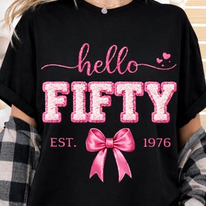 Puede incluir: Camiseta negra con la frase "hello FIFTY" en rosa y un lazo rosa. La palabra "FIFTY" está en una fuente texturizada y en bloque. El texto "EST. 1976" está debajo. Diseño temático de cumpleaños.