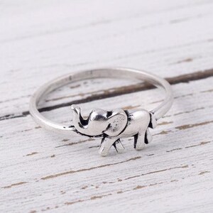 Könnte beinhalten: Silberner Ring mit einem kleinen Elefanten-Anhänger. Der Elefant hat detaillierte Stoßzähne und einen strukturierten Körper. Der Ring liegt auf einer weißen Holzoberfläche.
