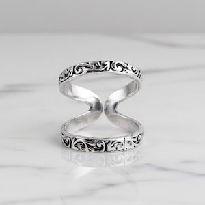 Könnte beinhalten: Ein silberner Doppel-Fingerring mit offenem Design. Der Ring hat ein kompliziertes, sich wiederholendes Scrollmuster um die Bänder. Der Ring wird auf einer weißen Marmoroberfläche mit seinem Spiegelbild gezeigt.