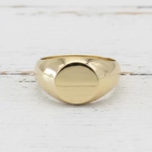 Könnte beinhalten: Ein goldener Siegelring mit glatter, polierter Oberfläche. Der Ring hat eine abgerundete, ovale Oberfläche und ein dickes Band. Der Ring ist vor einem weißen Holzhintergrund platziert.