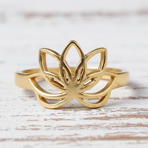 Könnte beinhalten: Ein goldfarbener Ring mit einem Lotusblumen-Design. Der Ring hat ein offenes Design mit einem floralen Motiv. Der Ring ist vor einem weißen Holzhintergrund platziert.