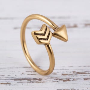 Könnte beinhalten: Ein goldfarbener Pfeilring. Der Ring hat ein Pfeildesign mit dreieckiger Pfeilspitze und dekorativer Befiederung. Der Ring ist vor einem weißen Holzhintergrund platziert.