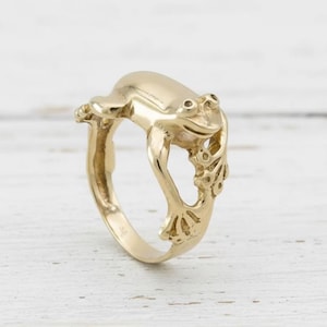 Könnte beinhalten: Ein goldfarbener Ring mit einem Frosch-Design. Der Frosch ist im Profil dargestellt, sein Körper wölbt sich über das Band des Rings. Der Ring hat ein detailliertes, strukturiertes Aussehen mit sichtbaren Augen und Gliedmaßen. Der Ring befindet sich auf einer weißen Holzoberfläche.