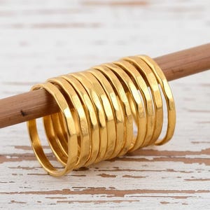 Könnte beinhalten: Eine Sammlung goldfarbener Ringe mit gehämmerten Texturen, auf einen Holzstab aufgefädelt. Die Ringe sind dünn und haben eine glänzende Oberfläche. Der Stab ist hellbraun, der Hintergrund ist eine weiße Holzoberfläche.