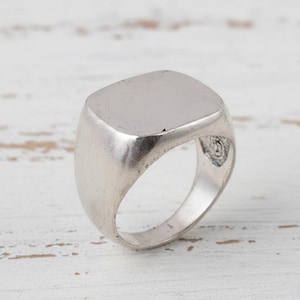 Könnte beinhalten: Ein silberfarbener Siegelring mit quadratischer Oberfläche. Der Ring hat eine glatte, polierte Oberfläche und ein klassisches Design. Der Ring ist wahrscheinlich aus Metall und für den täglichen Gebrauch geeignet.