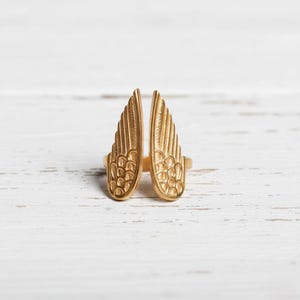 May include: A gold-colored ring featuring a design of two open wings. The wings have detailed feather patterns and are positioned upright. The ring is set against a white wooden surface, highlighting the jewelry's metallic sheen.