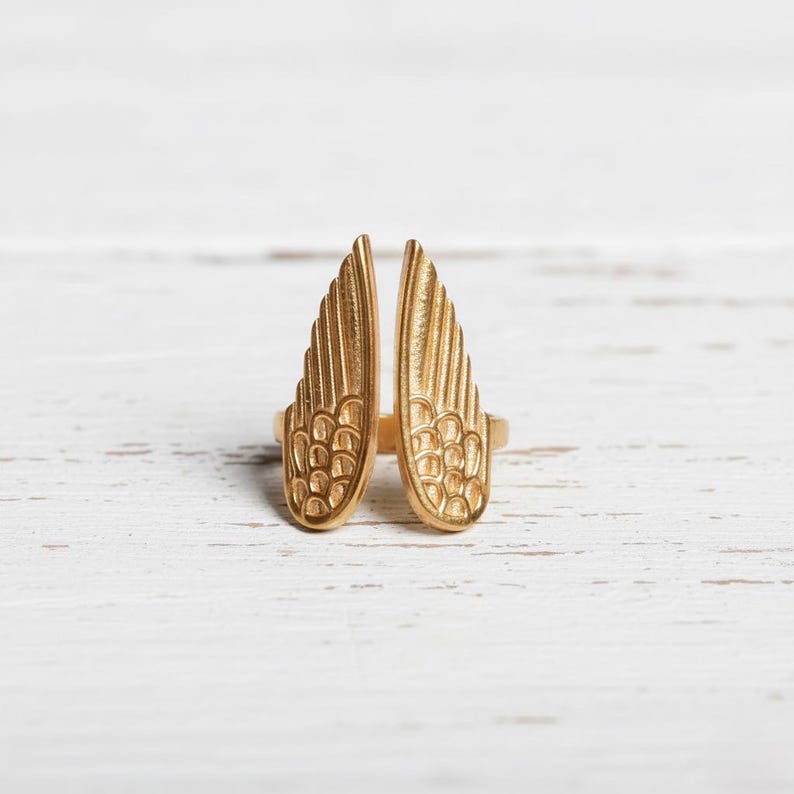 May include: A gold-colored ring featuring a design of two open wings. The wings have detailed feather patterns and are positioned upright. The ring is set against a white wooden surface, highlighting the jewelry's metallic sheen.