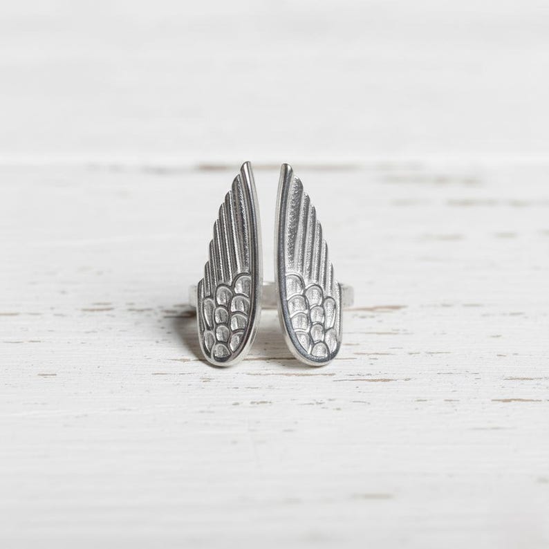 May include: A silver-colored ring featuring a split wing design. Each wing is detailed with feather-like patterns, creating a textured appearance. The ring is set against a white wooden surface, highlighting the jewelry's metallic sheen.