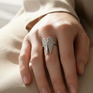 May include: A silver-toned ring featuring an open wing design. The ring has two detailed wings, one on each side, with a textured surface. The ring is worn on a finger, showcasing the jewelry's design and craftsmanship. The ring is a piece of fashion jewelry.