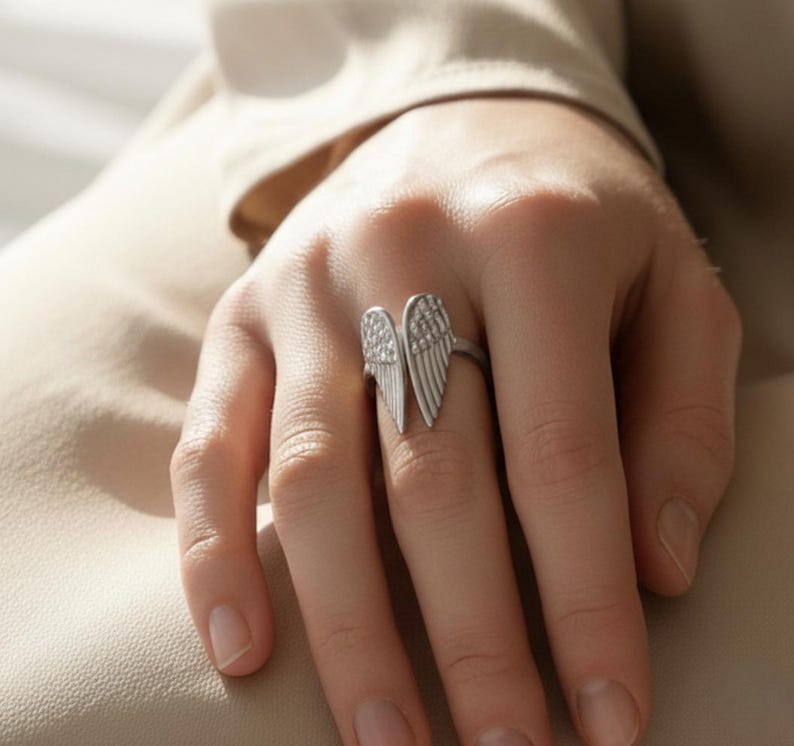 May include: A silver-toned ring featuring an open wing design. The ring has two detailed wings, one on each side, with a textured surface. The ring is worn on a finger, showcasing the jewelry's design and craftsmanship. The ring is a piece of fashion jewelry.