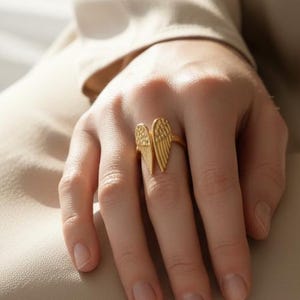 May include: A gold-colored ring featuring a stylized wing design. The ring's wings are detailed with textured lines, creating a feather-like appearance. The ring is worn on a finger, showcasing its design and craftsmanship.