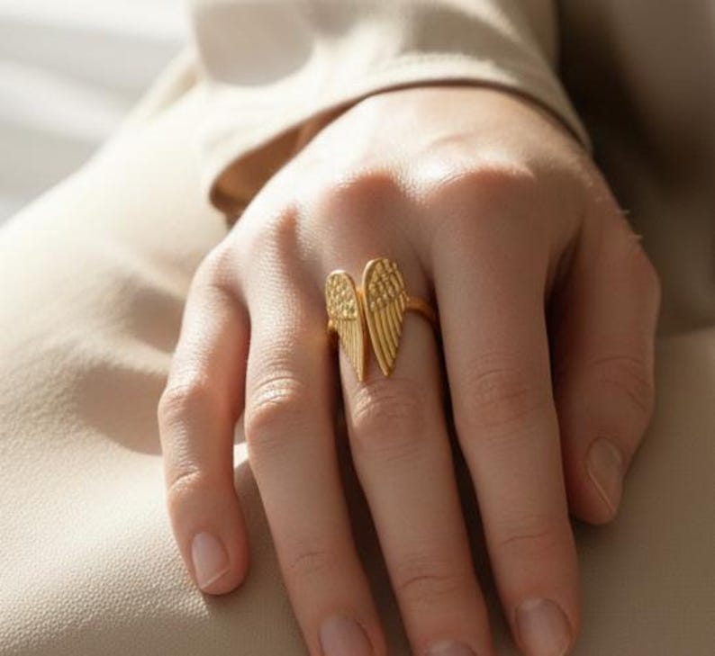 May include: A gold-colored ring featuring a stylized wing design. The ring's wings are detailed with textured lines, creating a feather-like appearance. The ring is worn on a finger, showcasing its design and craftsmanship.