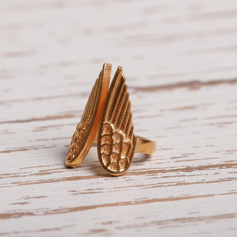 May include: A gold-colored ring featuring a wing design. The ring has two detailed wings that extend upwards. The wings have a textured surface with a feather-like pattern. The ring is set against a light-colored wooden surface.